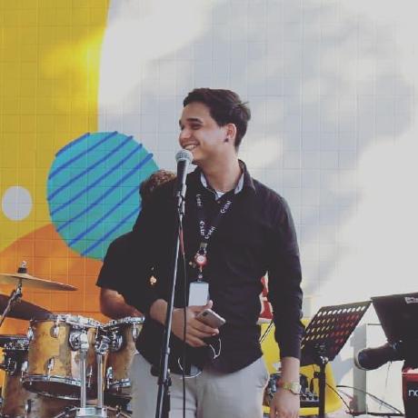 Foto de Víctor Martínez, se identifica como el/dele, é uma menino braco, com cabelo curto preto, camisa social azul escuro, e calça cinza, está parado em frente um microfone, com un fundo de cores amarelo e azul